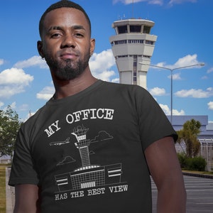 Può includere: Una maglietta nera con una grafica bianca di una torre di controllo aeroportuale e un aereo. Il testo "My Office Has The Best View" è stampato sulla maglietta.