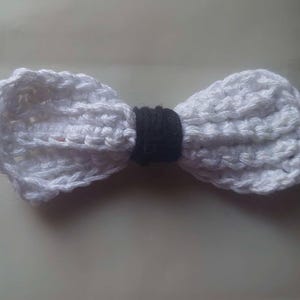 May include: A white crocheted bow with a black center. The bow is made of yarn and is a handmade craft.