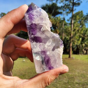May include: A purple and white fluorite crystal point with iridescent accents. The crystal is in the shape of a pyramid and has a rough, natural texture.