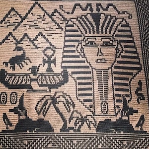 以下が含まれることがあります： エジプトのファラオが頭飾りをかぶった茶色と黒の画像が描かれた、かぎ針編みの壁掛け。背景には山と砂漠の風景が描かれている。