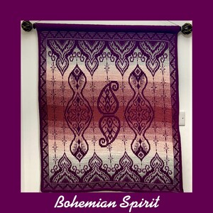 Op de afbeelding: Een paarse, roze en witte gehaakte wandhanger met een geometrisch patroon. De hanger hangt aan een witte muur met een houten stok aan de bovenkant. De tekst "Bohemian Spirit" staat onder de hanger.