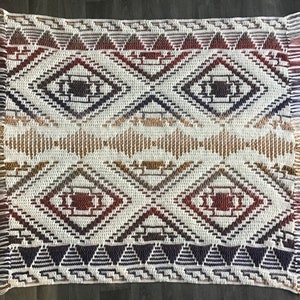 Könnte beinhalten: Ein weißer gewebter Teppich mit einem geometrischen Muster in Braun-, Rot- und Lilatönen. Der Teppich hat an allen vier Seiten Fransen.