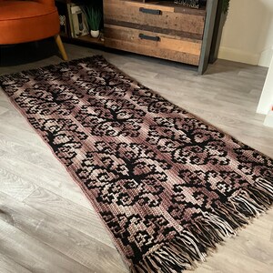May include: A brown and black crocheted rug with fringe on the edges. The rug has a geometric pattern and is laid out on a wood floor.