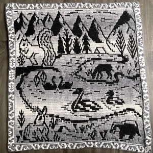 May include: A black and white crochet blanket with a woodland scene featuring trees, animals, and a river. The blanket has a gray background with black and white details.