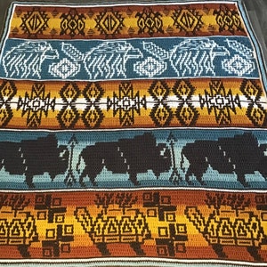 Op de afbeelding: Een gehaakt deken met een Native American ontwerp in de kleuren bruin, blauw en geel. Het deken heeft een geometrisch patroon met afbeeldingen van buffels en andere dieren.