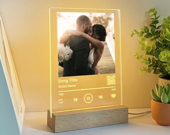 May include: A personalized LED light featuring a wedding photo, song title, and artist name. The clear acrylic panel is illuminated by a warm glow, set on a wooden base. The design includes a music player interface with play/pause and skip controls, and a QR code.