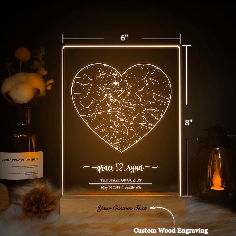 May include: A white LED night light with a wooden base. The light features a heart-shaped design with a constellation pattern inside. The text "grace ryan" is engraved below the heart, along with the date "May 30 2018" and the location "Seattle WA". The text "Your Custom Text" is engraved on the wooden base.