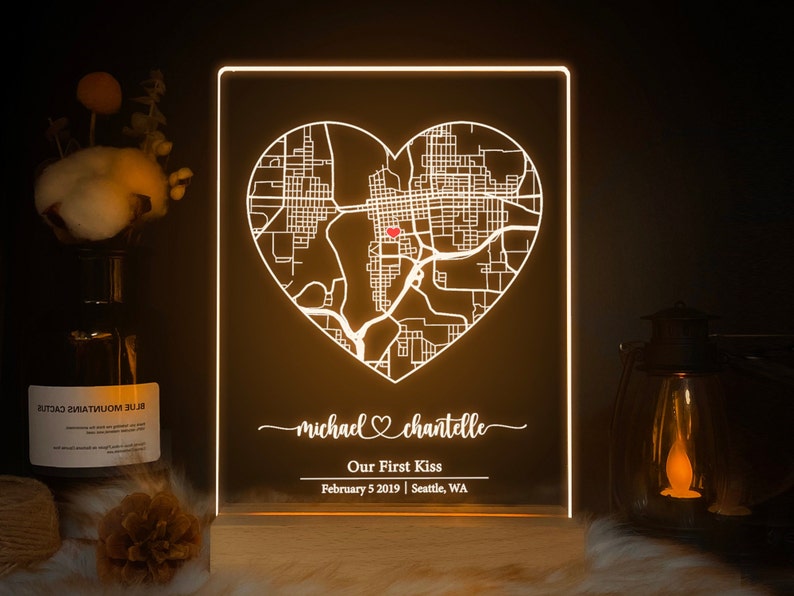 Pu&ograve; includere: Una mappa a forma di cuore bianca illuminata con un punto rosso al centro. La mappa &egrave; su uno sfondo nero con il testo "michael chantelle" in corsivo, seguito da "Our First Kiss" e "February 5 2019 | Seattle, WA".