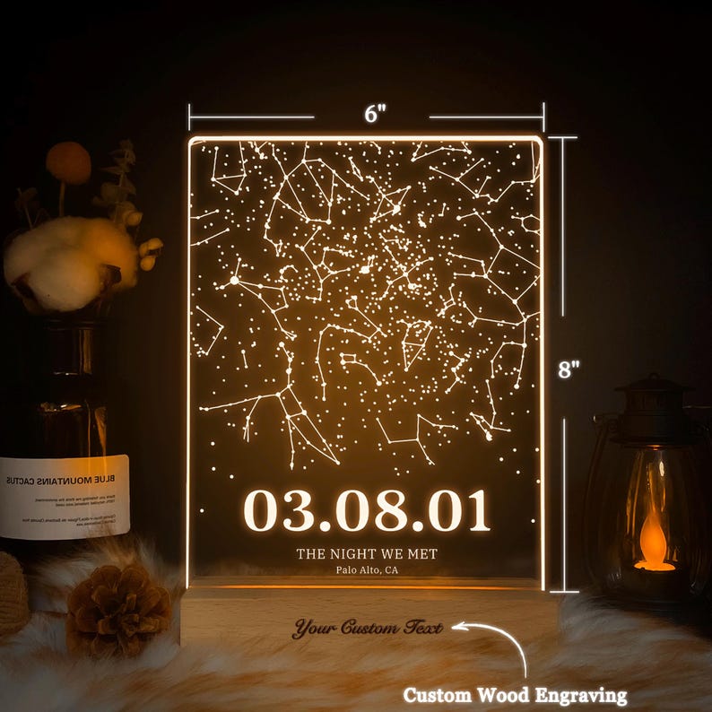 Puede incluir: Una luz de constelaci&oacute;n de cielo nocturno personalizada con una base de madera. La luz presenta un dise&ntilde;o de constelaci&oacute;n con una fecha, "03.08.01", y el texto "The Night We Met" con la ubicaci&oacute;n "Palo Alto, CA". La base tiene espacio para un mensaje grabado personalizado.