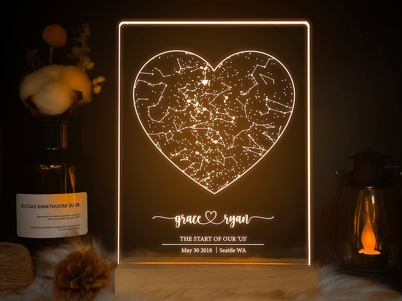 May include: A white LED night light shaped like a heart with a constellation design inside. The light has a wooden base and displays the names "grace ryan" and the text "THE START OF OUR 'US' May 30 2018 | Seattle WA".