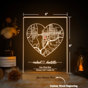 Pu&ograve; includere: Una base in legno incisa su misura con un pannello in acrilico bianco che presenta una mappa della citt&agrave; a forma di cuore con un punto rosso che segna una posizione. Il testo "michael chantelle" &egrave; scritto in corsivo con un simbolo di cuore tra i nomi. Il testo "Our First Kiss" &egrave; sotto i nomi, seguito dalla data "February 5 2019" e dalla posizione "Seattle, WA". La base ha il testo "Your Custom Text" e "Custom Wood Engraving".