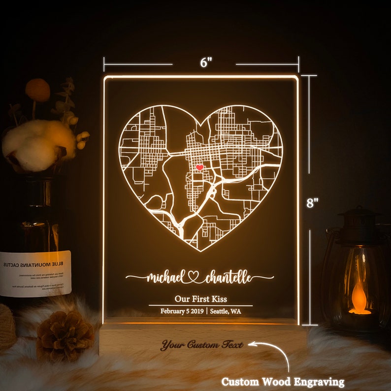 Pu&ograve; includere: Una base in legno incisa su misura con un pannello in acrilico bianco che presenta una mappa della citt&agrave; a forma di cuore con un punto rosso che segna una posizione. Il testo "michael chantelle" &egrave; scritto in corsivo con un simbolo di cuore tra i nomi. Il testo "Our First Kiss" &egrave; sotto i nomi, seguito dalla data "February 5 2019" e dalla posizione "Seattle, WA". La base ha il testo "Your Custom Text" e "Custom Wood Engraving".