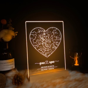 May include: A white LED light with a wooden base features a constellation-filled heart design. The text "grace ryan" and "THE START OF OUR US" is displayed below the heart, along with the date "May 30 2018" and location "Seattle WA".