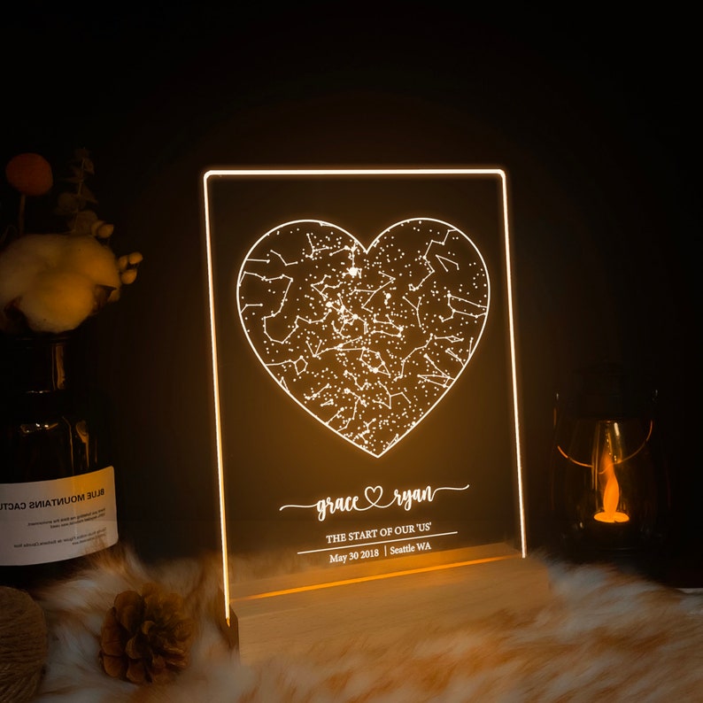 May include: A white LED light with a wooden base features a constellation-filled heart design. The text "grace ryan" and "THE START OF OUR US" is displayed below the heart, along with the date "May 30 2018" and location "Seattle WA".