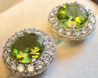 May include: Two round earrings with a central, faceted, light green gemstone surrounded by a halo of smaller, clear gemstones. The earrings have a silver-colored metal setting.