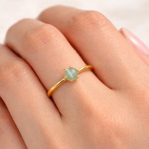 May include: A thin gold band ring featuring a single, small, round, pale green cabochon gemstone set in a six-prong setting. The smooth, polished metal band has a simple, slender design that encircles the finger with a minimalist aesthetic.