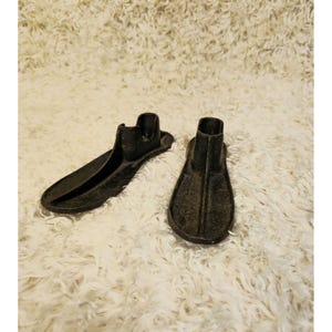 May include: Two antique black cast iron shoe lasts, one with a long, narrow shape and the other with a more rounded toe. The lasts are displayed on a fluffy, white textured surface.