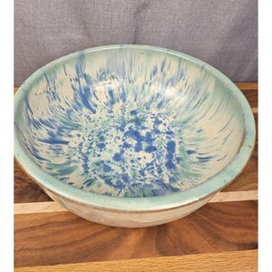 Könnte beinhalten: Eine runde Keramikschale mit einem gesprenkelten Glasurmuster in Blau-, Türkis- und Weißtönen. Das Innere zeigt ein lebendiges, strahlenförmiges Spritzdesign, das in einen hellfarbigen Rand und einen hellen äußeren Boden übergeht.