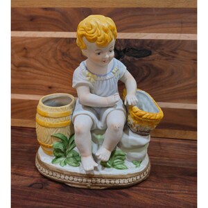 Könnte beinhalten: Porzellanfigur eines Kindes mit blonden Haaren, das ein hellblaues Outfit trägt und neben einem kleinen Fass und einem Korb sitzt. Die Figur steht auf einem dekorativen Sockel mit grünen Blättern.