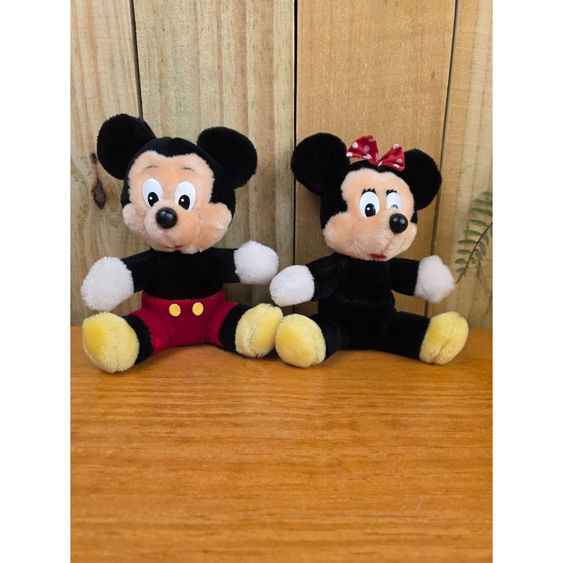 K&ouml;nnte beinhalten: Zwei Pl&uuml;schpuppen von Mickey Mouse und Minnie Mouse. Mickey tr&auml;gt rote Shorts und gelbe Schuhe. Minnie hat eine rote Schleife und tr&auml;gt ein schwarzes Kleid. Beide Puppen haben schwarze Ohren und sitzen auf einer Holzoberfl&auml;che.