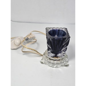 Puede incluir: Una pequeña lámpara de vidrio transparente con una pantalla de vidrio morado. La lámpara tiene un cable con un interruptor y una base de vidrio transparente con un diseño decorativo.