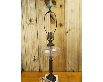 May include: Vintage table lamp with a clear glass globe and a dark metal base. The lamp features a teal blue orb finial and a curved metal shade holder. The lamp is on a white square base and has a white electrical cord.