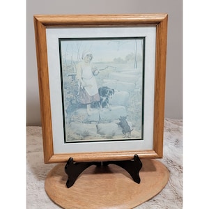 May include: A framed print of a girl in a white dress and a red apron walking with two dogs on a stone path. The print is in a wooden frame with a white mat.