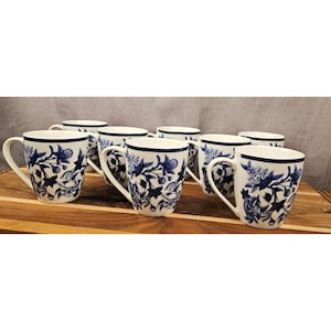 May include: A set of seven white ceramic mugs featuring a dark blue floral pattern. Each mug has a rounded handle and a consistent design with blue botanical illustrations wrapping around the exterior surface of the white porcelain drinkware.