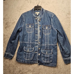 May include: A medium-wash blue denim jacket featuring a mandarin collar, a full-length front button closure, two chest pockets with buttoned flaps, and two lower patch pockets with buttoned flaps. The jacket has long sleeves and a structured fit.