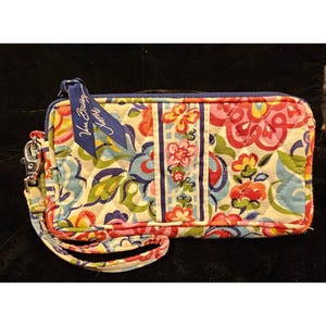 May include: A rectangular quilted fabric wristlet wallet featuring a multi-colored floral and butterfly pattern in shades of pink, blue, yellow, and green. A blue fabric tag with the text Vera Bradley is attached to the silver metal zipper pull.