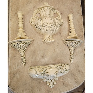 Puede incluir: Un conjunto de tres piezas de decoración de pared de cerámica blanca con diseños ornamentados. El conjunto incluye una pieza grande en forma de urna con tapa y dos portavelas más pequeños con estantes.