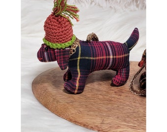 May include: A small, red and black plaid dachshund ornament with a green and brown crocheted hat. The ornament is sitting on a wooden surface.