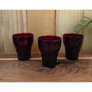 May include: Three dark red drinking glasses with a textured base are arranged on a wooden surface. The glasses have a rounded shape and a deep red color, perfect for serving beverages. The background is a wooden wall.