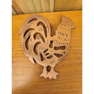 May include: Copper rooster-shaped trivet with a cutout design. The trivet has a ring for hanging and is made of copper-colored metal. The rooster's tail feathers and body are detailed with cut-out shapes. The trivet is placed on a wooden surface.