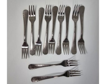 May include: A collection of stainless steel forks with a hammered texture on the handles. The forks are arranged on a white surface, showcasing their tines and overall design. The set includes various forks for dining.