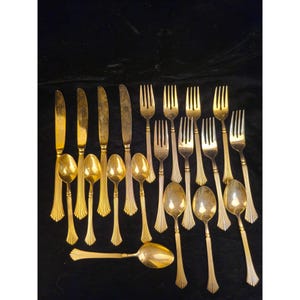 May include: A set of gold-coloured cutlery, including knives, forks, and spoons, displayed on a dark background. The silverware features a decorative, ridged pattern on the handles. The arrangement suggests a formal dining setting.