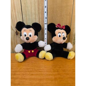 K&ouml;nnte beinhalten: Zwei Pl&uuml;schfiguren von Mickey und Minnie Mouse. Mickey tr&auml;gt rote Shorts und gelbe Schuhe. Minnie hat eine rote Schleife. Beide haben schwarze Ohren, schwarze K&ouml;rper und wei&szlig;e Handschuhe. Etwa 20 cm hoch.