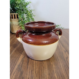 Puede incluir: Un macetero de cerámica marrón y blanco con dos asas. El macetero tiene un esmalte marrón en la parte superior e inferior con una banda blanca en el medio.