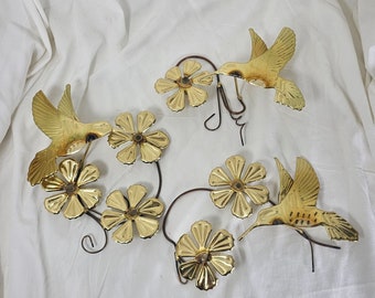 Pode incluir: Arte de parede de metal dourado com três beija-flores e seis flores. Os beija-flores estão em voo e as flores são estilizadas com cinco pétalas.