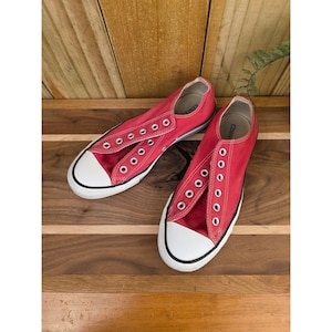 Op de afbeelding: Een paar rode canvas sneakers met witte rubberen neuzen en zolen. De sneakers hebben witte veterogen en worden gepresenteerd op een houten oppervlak. Een klassieke, casual stijl.