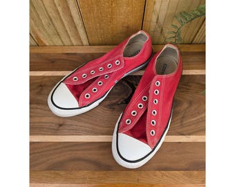 Puede incluir: Un par de zapatillas rojas de lona con punteras y suelas de goma blancas. Las zapatillas tienen ojales blancos para los cordones y se exhiben sobre una superficie de madera. Un estilo clásico e informal.