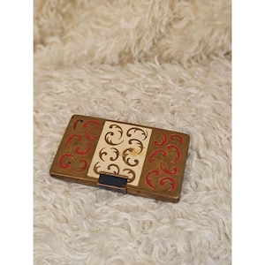 Op de afbeelding: Een rechthoekige, antiek messingkleurige doos met een crèmekleurig middenpaneel en rode en crèmekleurige krullen. De doos heeft een zwarte sluiting en staat tegen een pluizige, witte achtergrond.