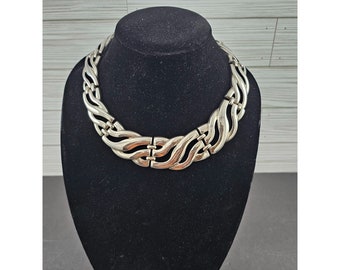 May include: A silver tone statement necklace with a thick, chunky chain design. The chain is made up of interlocking links that create a wavy pattern.
