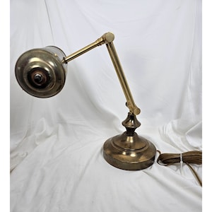 Puede incluir: Una lámpara de escritorio de latón con un brazo ajustable y una pantalla cilíndrica. La lámpara tiene una base redonda y un cable con un enchufe.