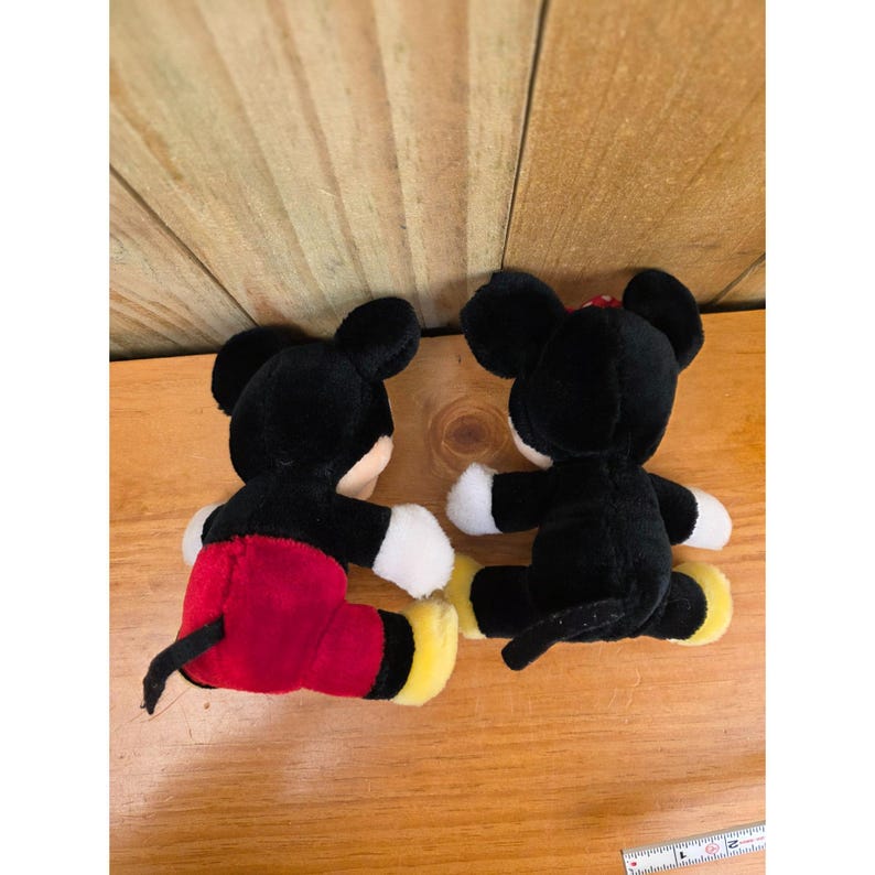 K&ouml;nnte beinhalten: Zwei Pl&uuml;schpuppen der Disney-Charaktere Micky Maus und Minnie Maus werden gezeigt. Micky tr&auml;gt rote Shorts, gelbe Schuhe und wei&szlig;e Handschuhe. Minnie hat eine rote Schleife, wei&szlig;e Handschuhe und gelbe Schuhe. Beide Puppen sind schwarz und wei&szlig;.