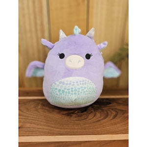 May include: A plush purple dragon toy with light blue wings and belly. The dragon has silver horns and a light blue crest. The belly features a light blue and white pattern. The dragon has black eyes and a white snout.