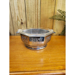 May include: A silver-colored, octagonal bowl with a clear, faceted rim. The bowl features decorative cut-out patterns and a pedestal base. Two small handles are on either side of the bowl. The bowl is likely used for serving or display.
