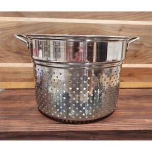 Puede incluir: Un colador de acero inoxidable con numerosos agujeros pequeños para escurrir. El colador tiene dos asas y un borde ancho. Se encuentra sobre una superficie de madera, mostrando su acabado metálico pulido. Esta herramienta de cocina está diseñada para lavar y colar alimentos.