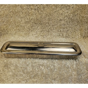 Puede incluir: Una fuente rectangular de acero inoxidable con tapa. La fuente tiene una superficie pulida y reflectante y un pequeño asa ovalada en la tapa. Los bordes son ligeramente redondeados.