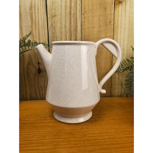 Pode incluir: Um jarro de cerâmica branca salpicada com bico e alça. O jarro tem uma base castanha clara e assenta numa superfície de madeira. O fundo apresenta tábuas de madeira e vegetação.